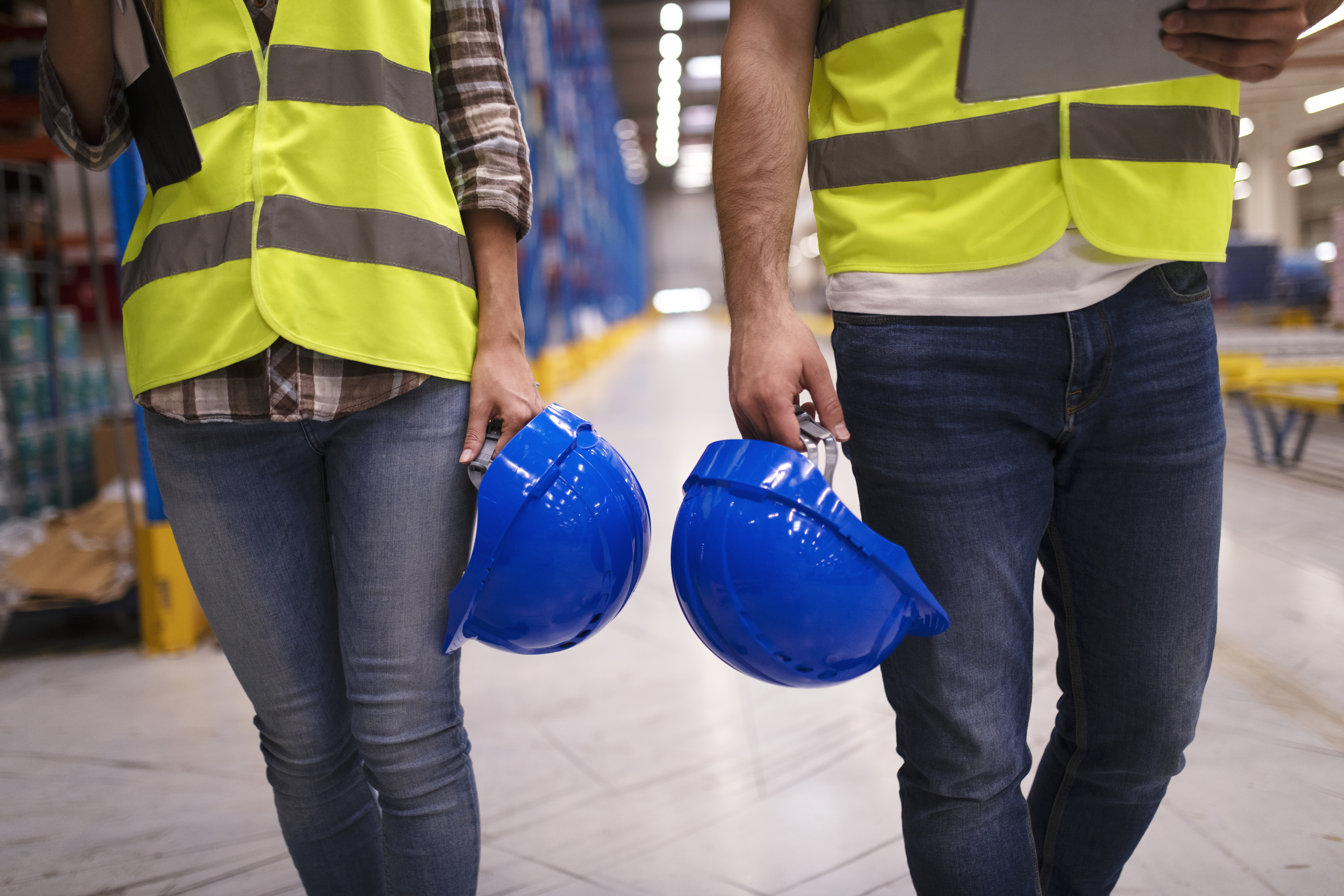 Two unrecognizable workers in reflective suit walking through warehouse and holding blue protective hardhats.